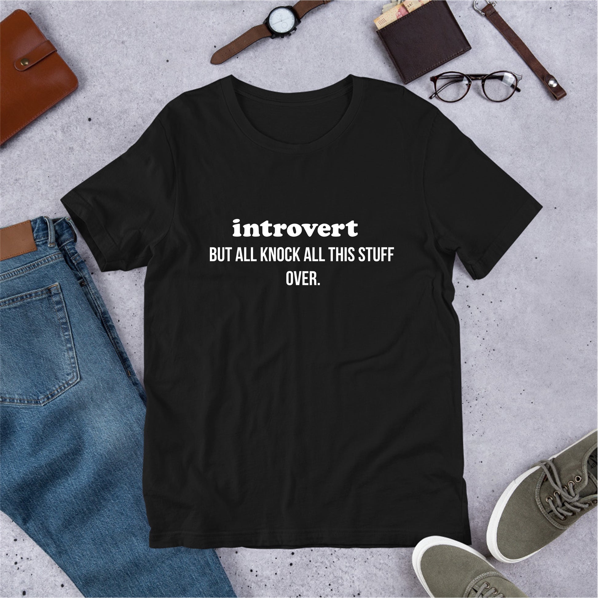 Black t-shirt with 'introvert BUT ALL KNOCK ALL THIS STUFF OVER.' text on a concrete surface with jeans, shoes, and accessories.