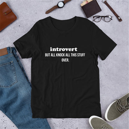 Black t-shirt with 'introvert BUT ALL KNOCK ALL THIS STUFF OVER.' text on a concrete surface with jeans, shoes, and accessories.