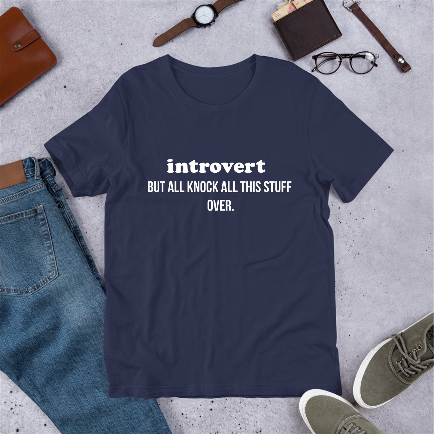 Navy blue t-shirt with 'introvert BUT ALL THIS STUFF OVER.' text on a gray surface with jeans, shoes, and accessories.