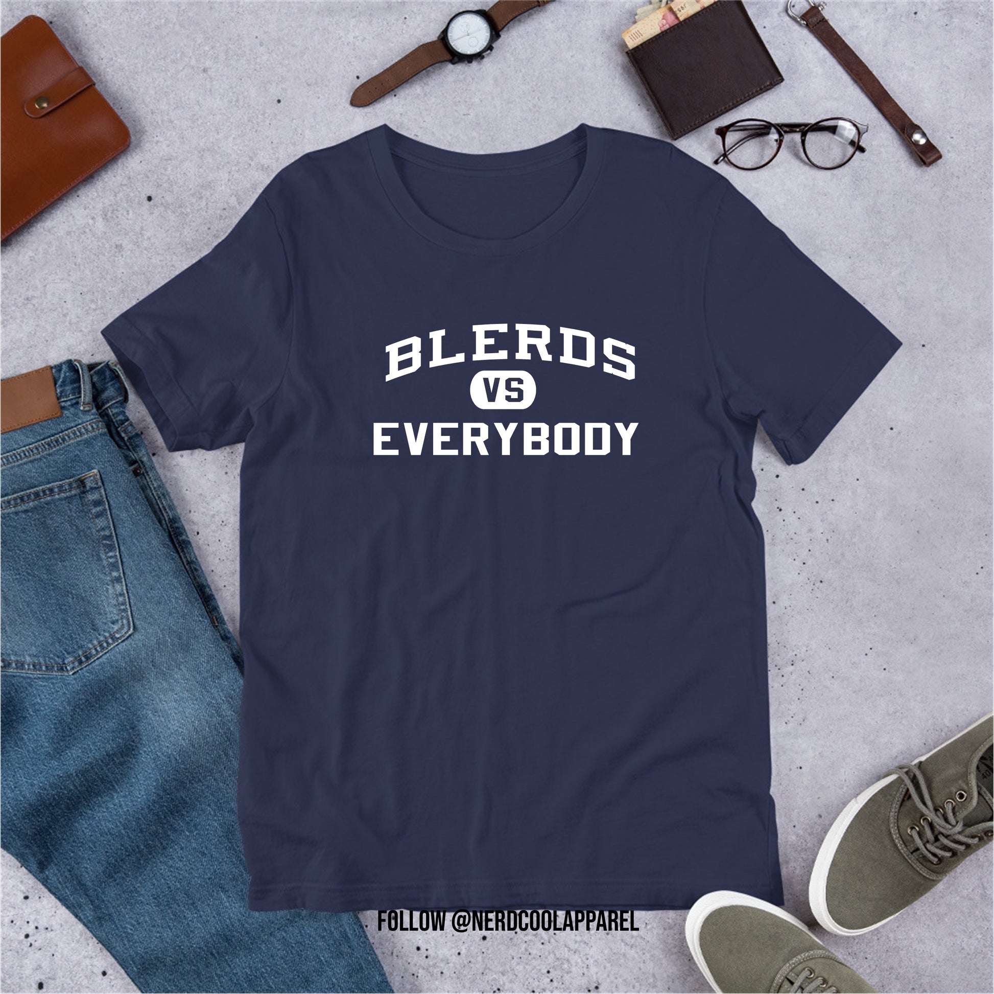 Navy blue t-shirt with 'BLERDS VS EVERYBODY' text on a gray surface with jeans, shoes, and accessories.