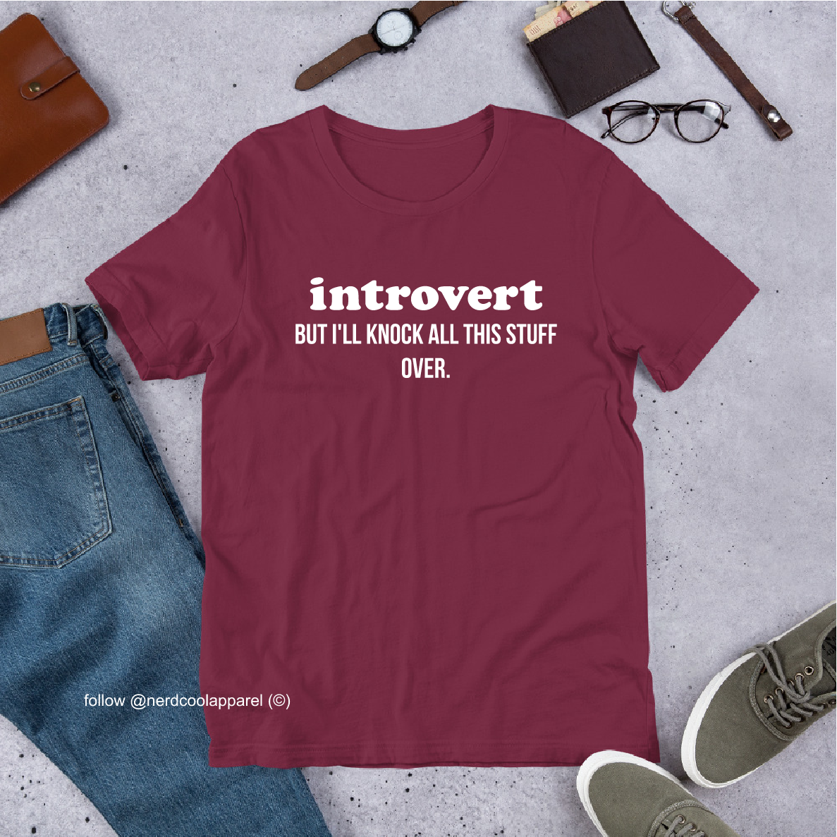 Maroon t-shirt with 'introvert BUT I'LL KNOCK ALL THIS STUFF OVER.' text, surrounded by jeans, shoes, and accessories on a gray surface.